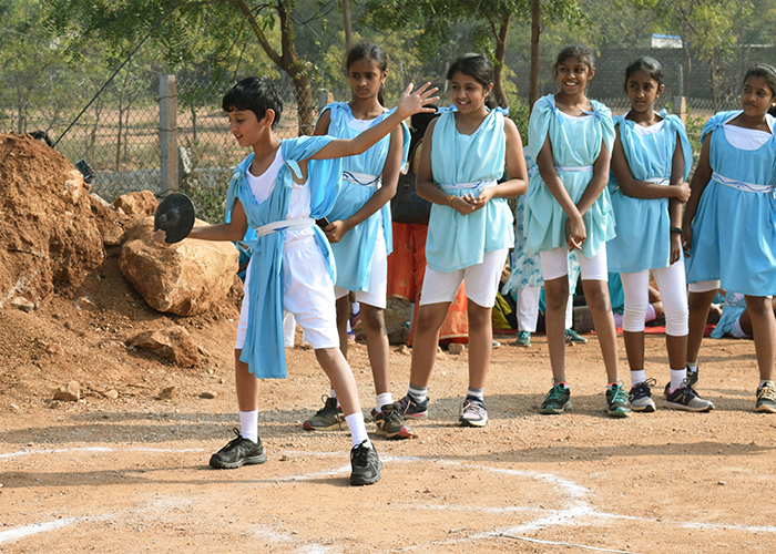 A middle school student dressed in traditional Greek garb engages in discus throw on a playground while classmates look on.