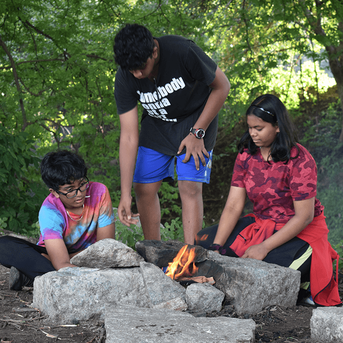Three students tending to a fire burning within a stone stove during outdoor activity.