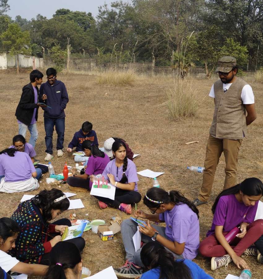 A cluster of students sit outdoors on the ground with painting materials while an instructor looks on.