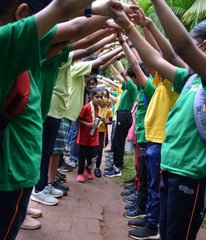 Older students stand in a row on either side of a path, raising hands to form a canopy under which younger students walk.