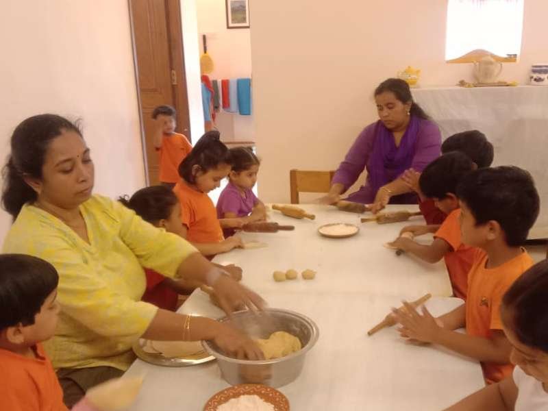 Students in a classroom seated around a long table rolling dough under the guidance of two teachers.