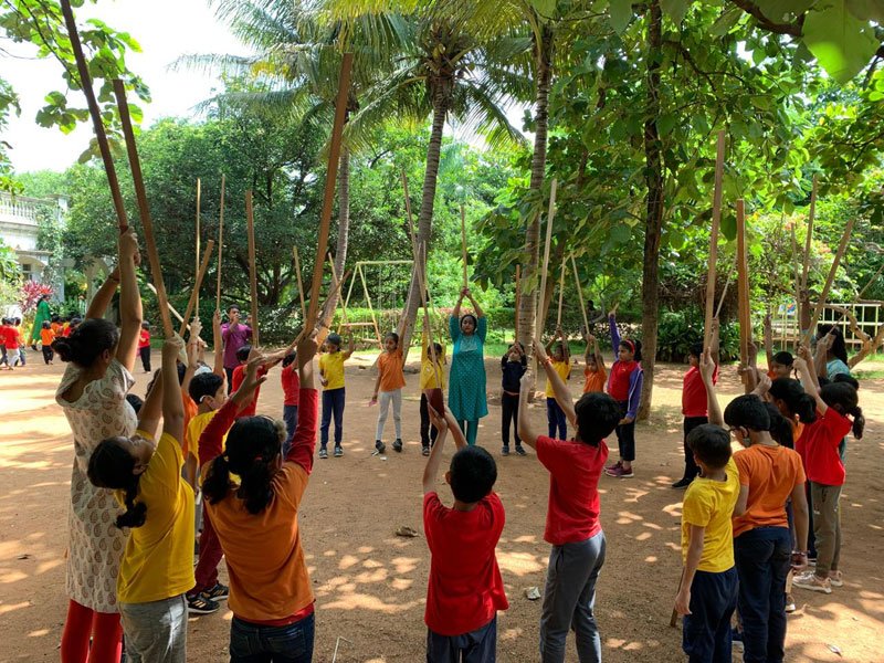 Students of a class gathered in a circle on a playground, waving sticks in the air in an outdoor lesson led by teachers.